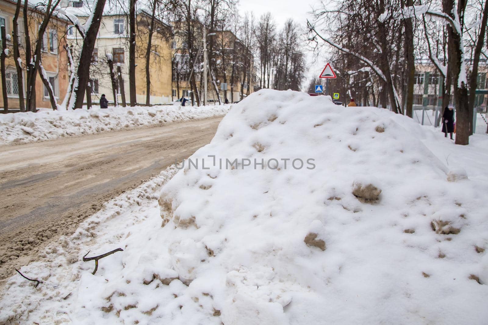 Snowy big snowdrift near the road against the backdrop of a city street. On the road lies dirty snow in high heaps. Urban winter landscape. Cloudy winter day, soft light.