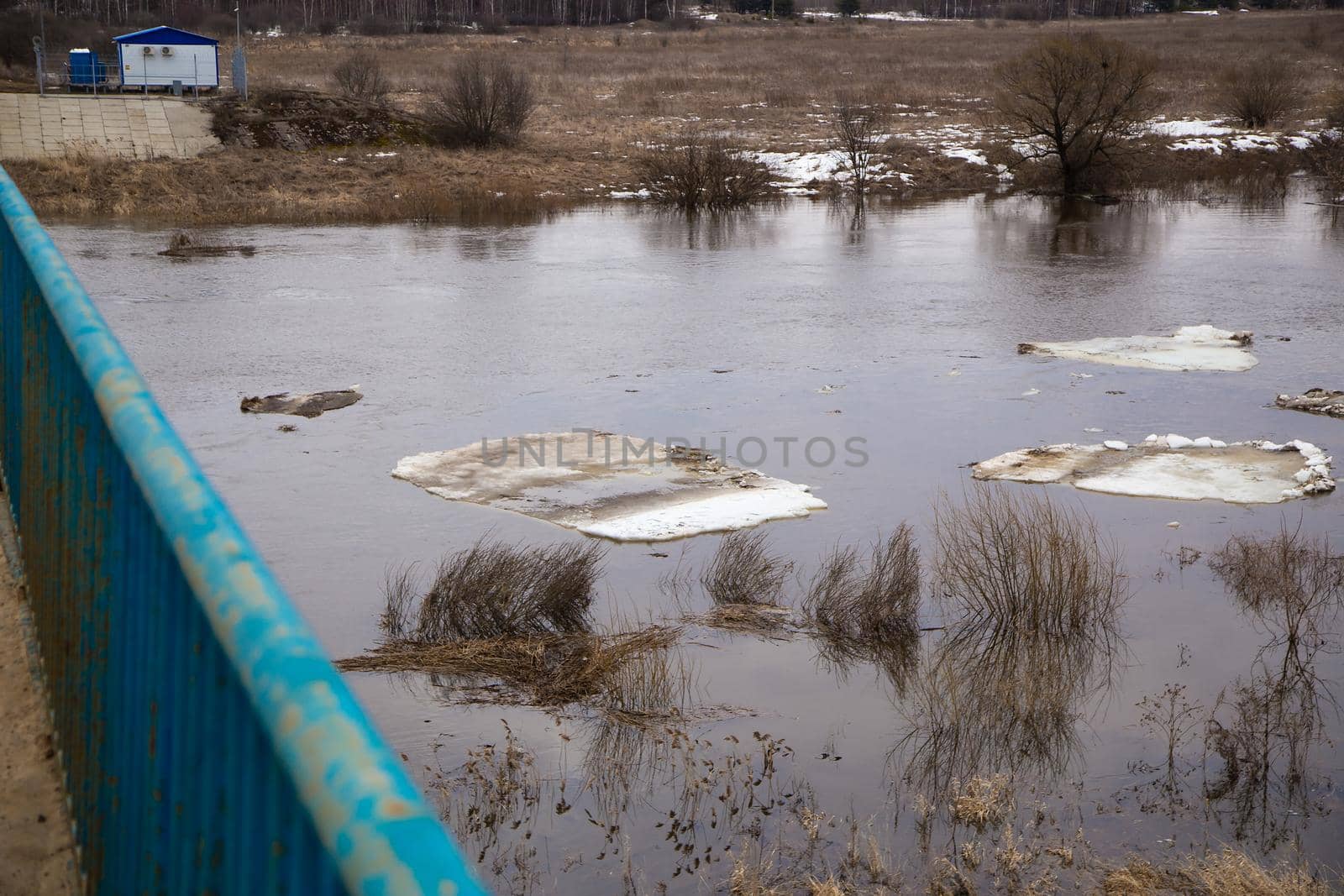 Dirty ice floes float down the river slowly. Spring, snow melts, dry grass all around, floods begin and the river overflows. Day, cloudy weather, soft warm light.