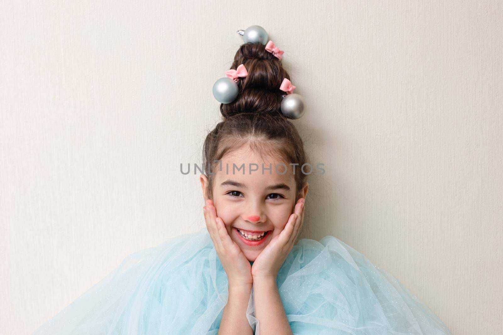 Portrait of a funny beautiful little girl in blue clothes, with a hairstyle in the form of a Christmas tree with a luminous garland, stands against the background of a light wall, pressed her palms to her cheeks