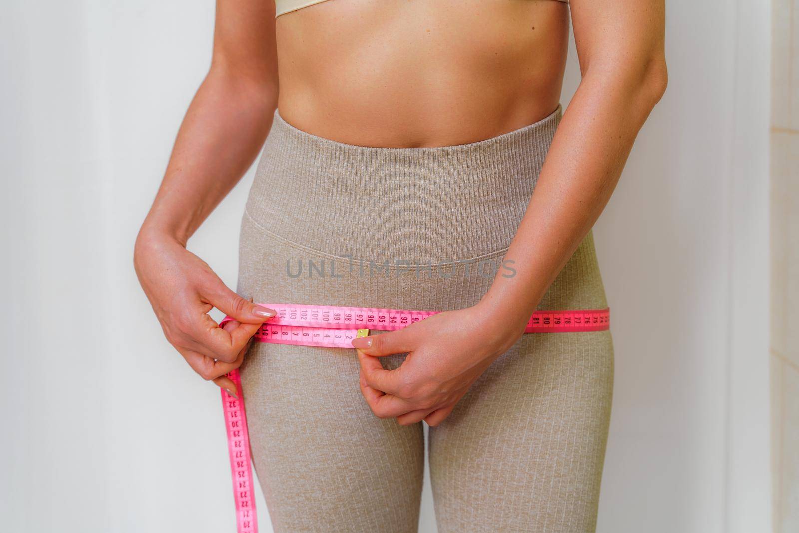 Cropped view of slim woman measuring hips with tape measure at home, close up. An unrecognizable European woman checks the result of a weight loss diet or liposuction indoors. Healthy lifestyle