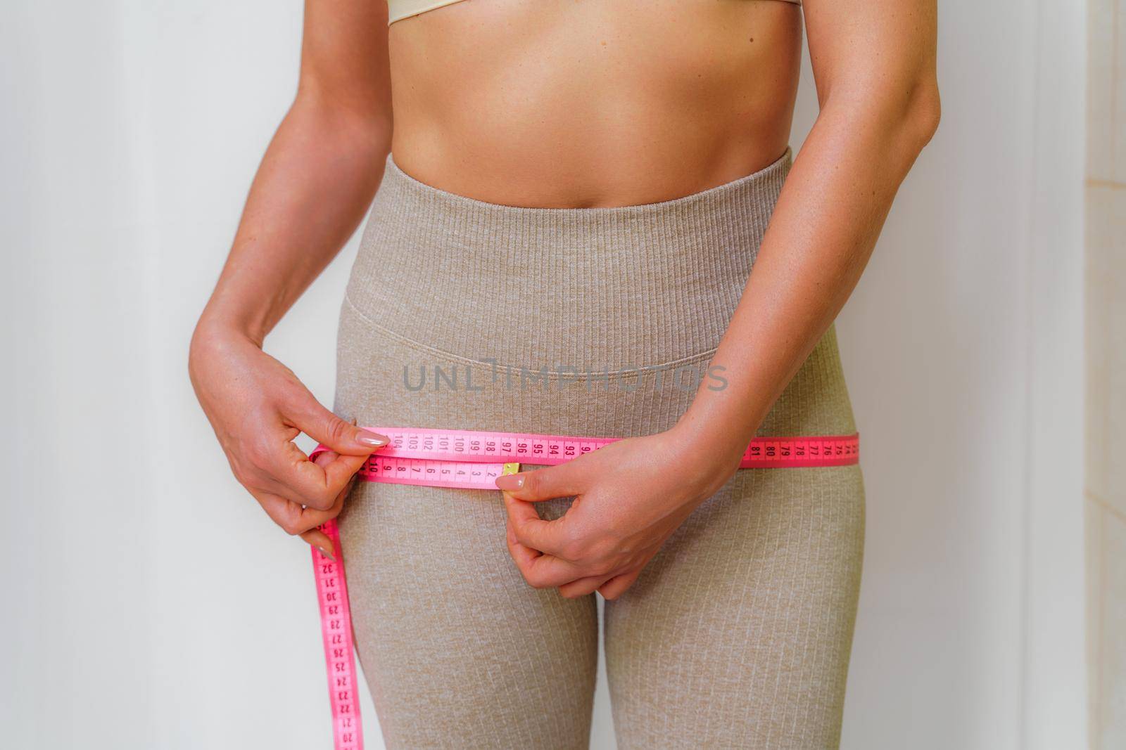 Cropped view of slim woman measuring hips with tape measure at home, close up. An unrecognizable European woman checks the result of a weight loss diet or liposuction indoors. Healthy lifestyle. by Matiunina