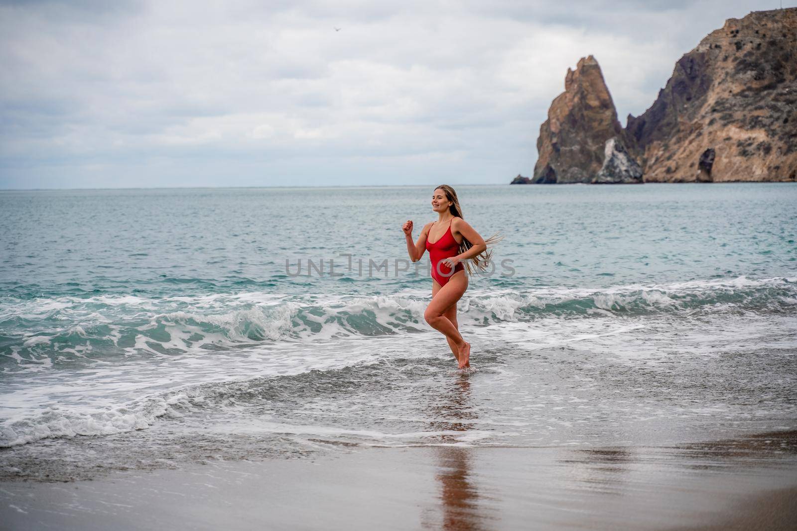 A beautiful and sexy brunette in a red swimsuit on a pebble beach, Running along the shore in the foam of the waves.