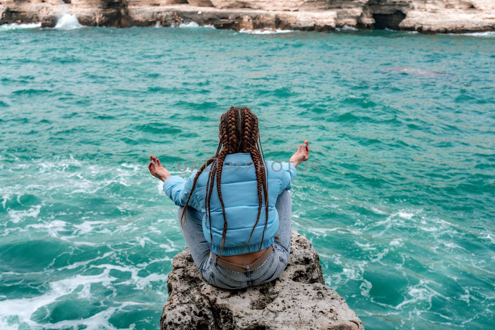 A woman in a blue jacket sits on a rock above a cliff above the sea, looking at the stormy ocean. Girl traveler rests, thinks, dreams, enjoys nature. Peace and calm landscape, windy weather