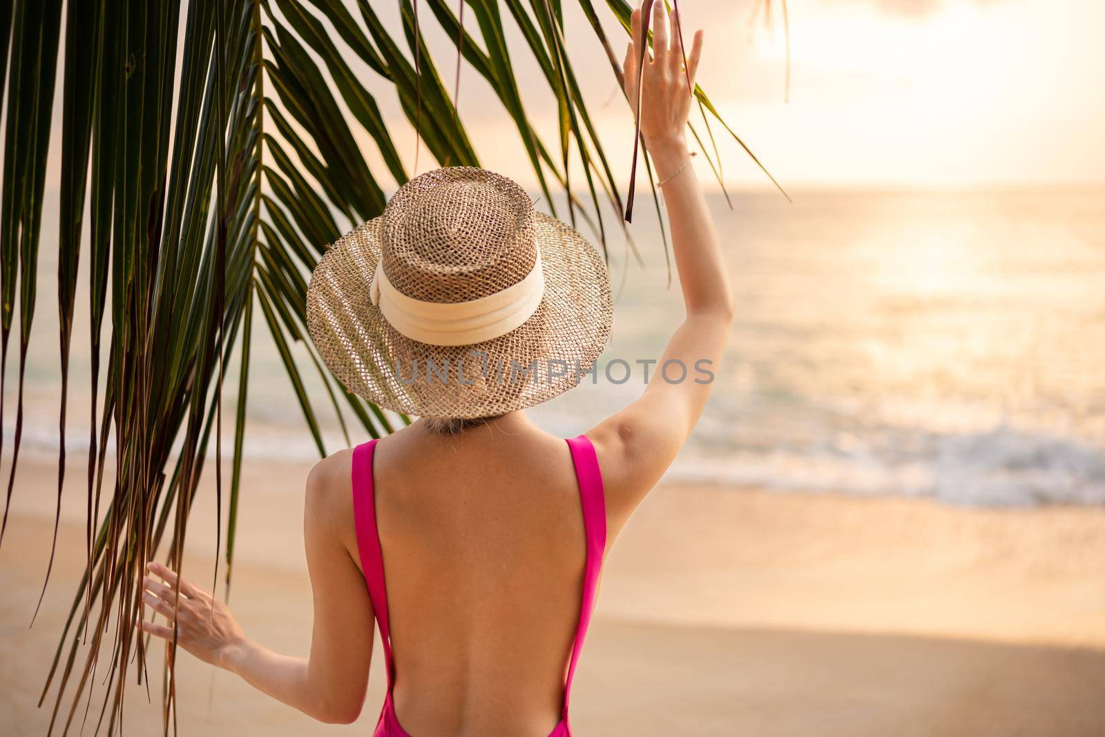 Woman wearing pink one piece swimsuit and straw hat enjoy romantic sunset moment under the coconut trees on tropical beach.