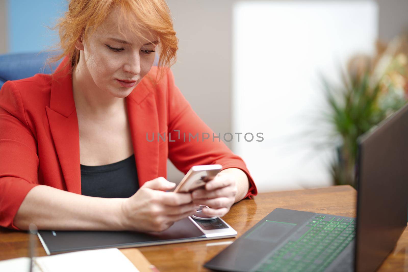 Redhead businesswoman using laptop and smartphone at the office by Novic