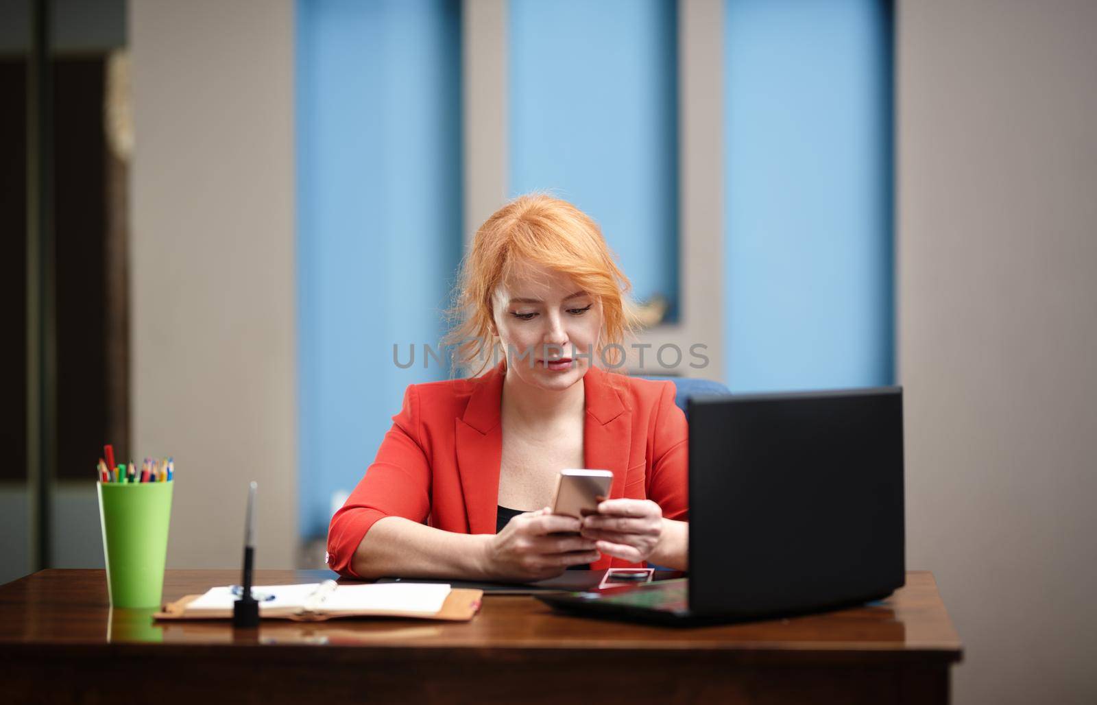 Redhead businesswoman using laptop and smartphone at the office by Novic