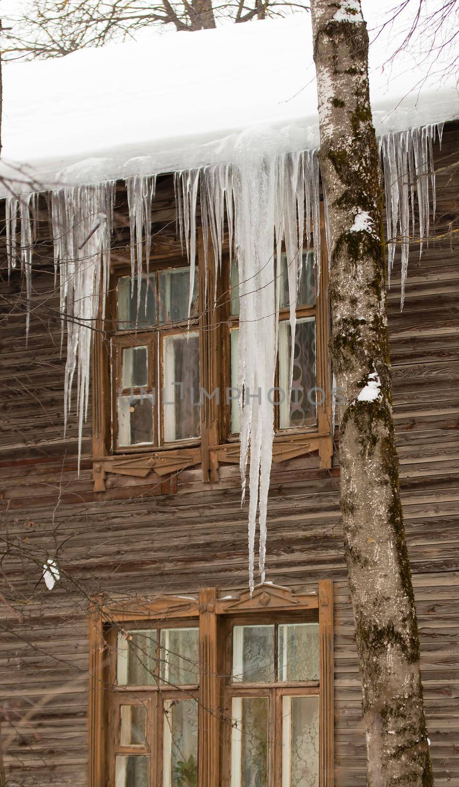 Fabulous transparent icicles hang on the edge of the roof. Against the background of the wooden wall of the old house. Large cascades, even beautiful rows. Cloudy winter day, soft light.