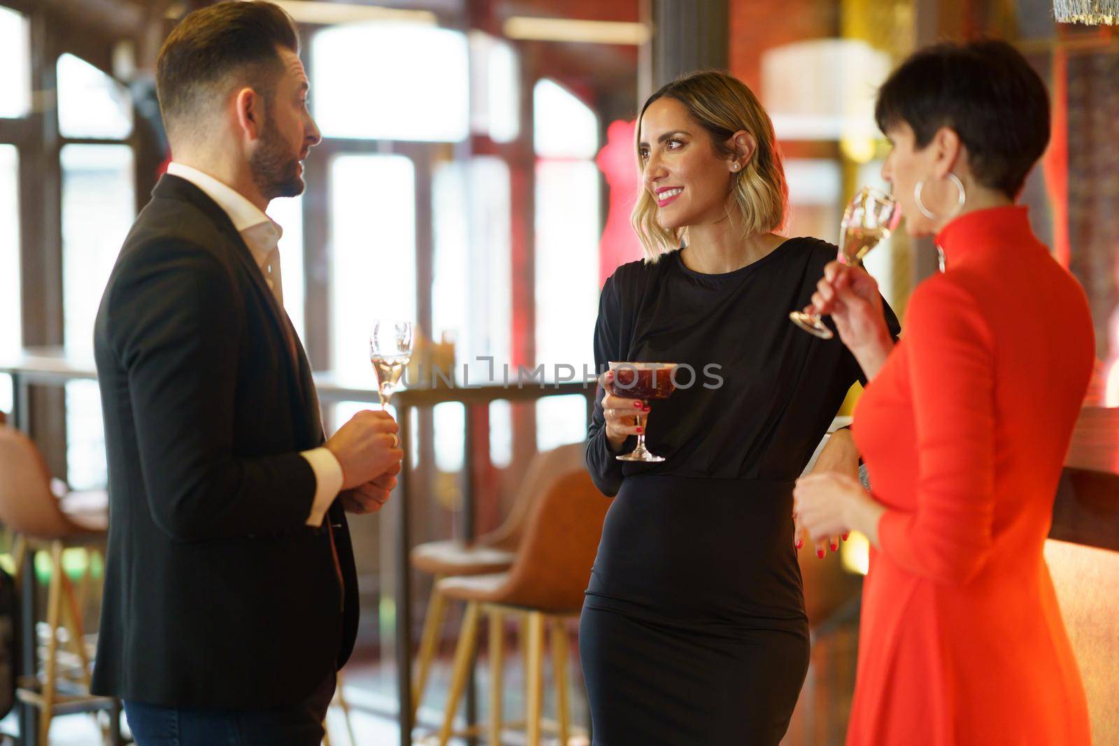 Group of well dressed friends drinking alcohol beverages and enjoying meeting while standing at wooden counter in bar with bartender