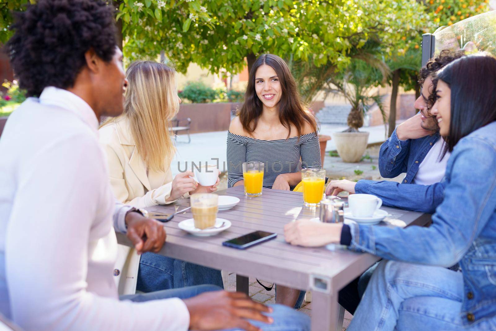 Multi-ethnic group of friends having a drink together in an outdoor bar. One of the men shows something on his smartphone to his friends.