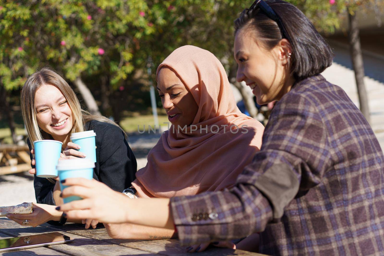 Cheerful young multiracial female coworkers laughing and communicating while having coffee break sitting at wooden table in park on sunny day