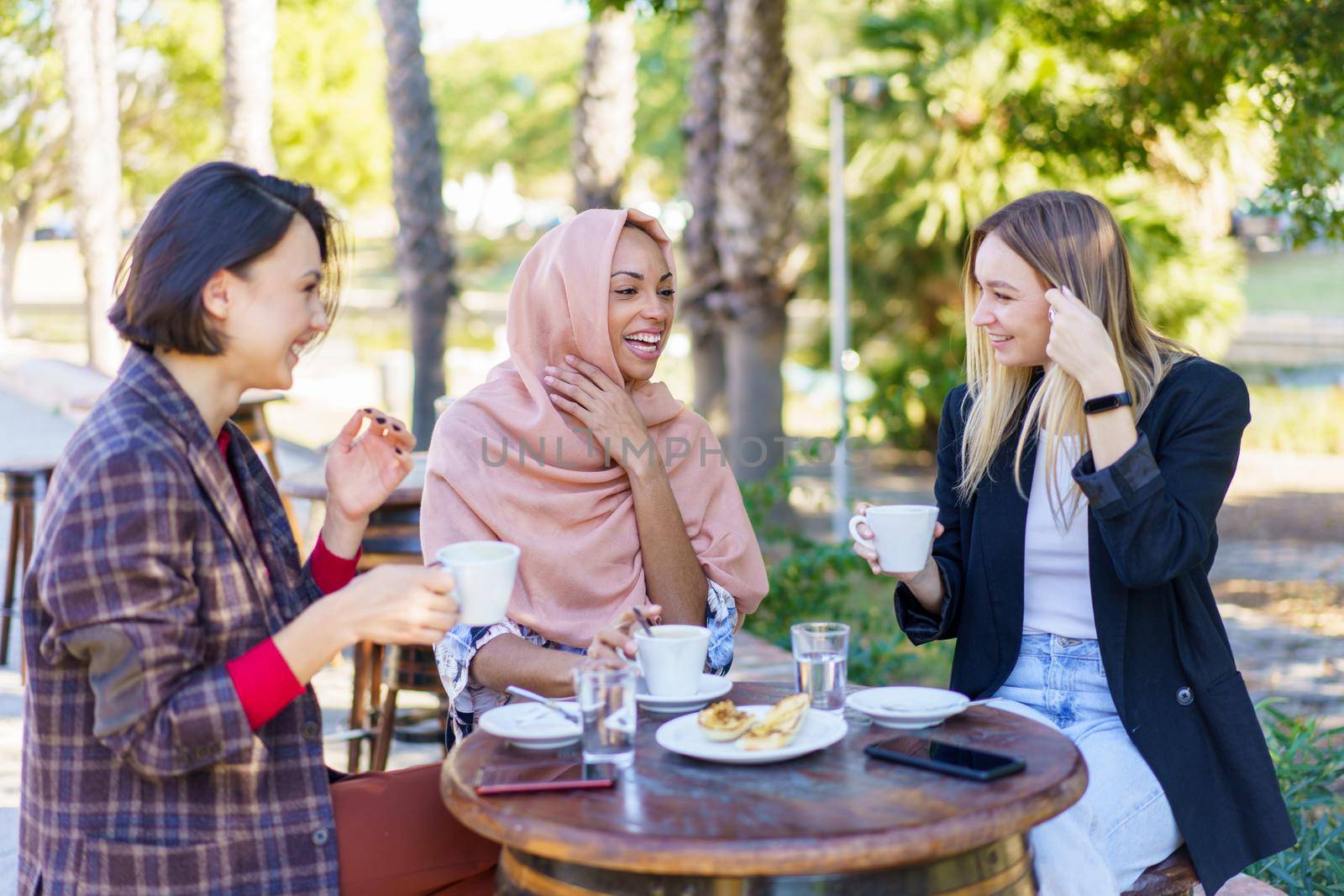 Group of smiling diverse female friends chatting while having coffee break at table on terrace of outdoor cafeteria in city