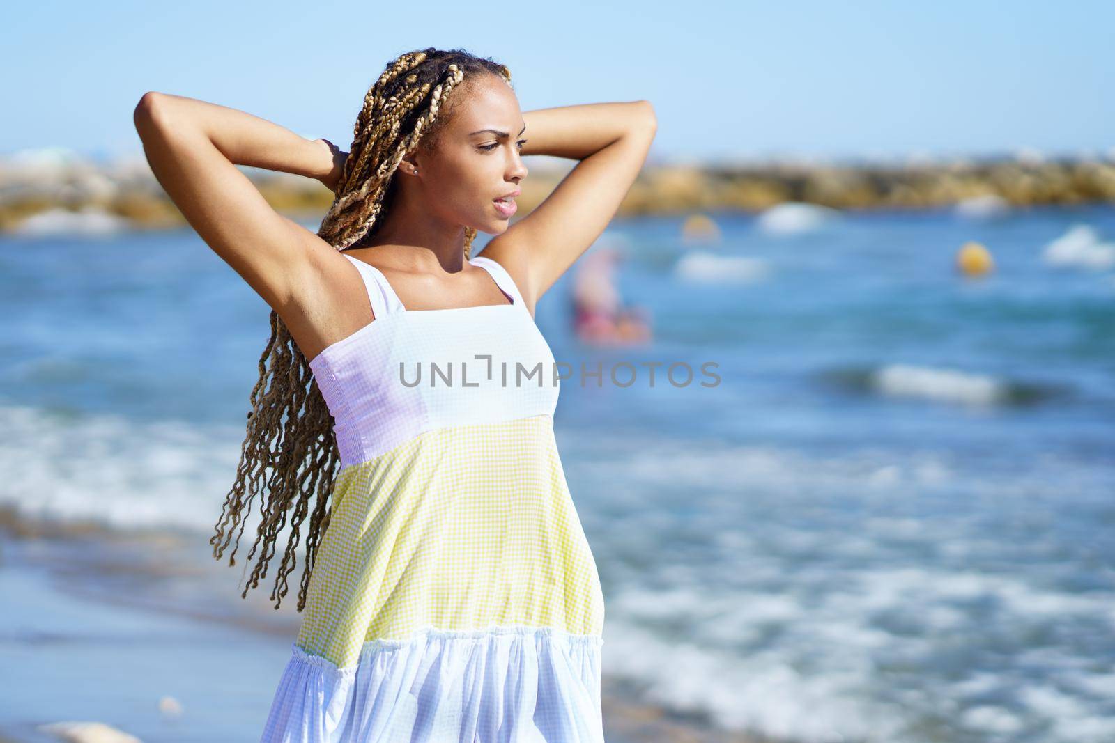 Black woman in summer dress with long braids holding hands behind head and looking away while relaxing on beach near waving sea on sunny day