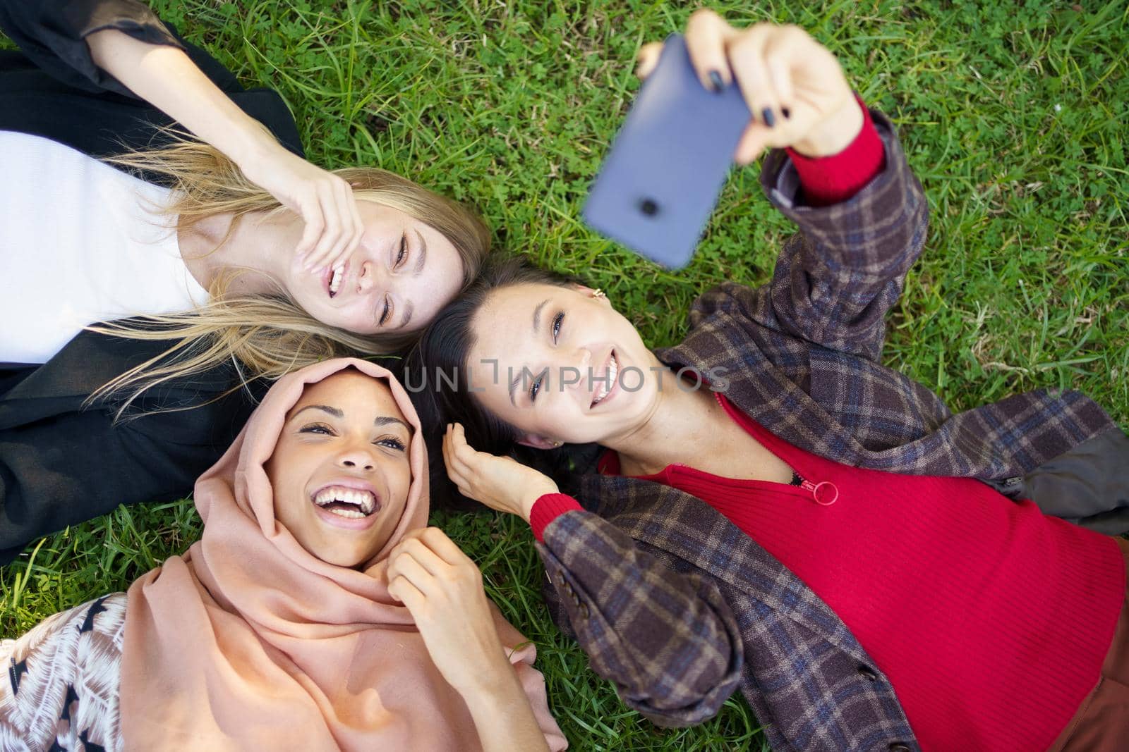 Top view of happy multiracial female friends lying on grass and taking self portrait on cellphone
