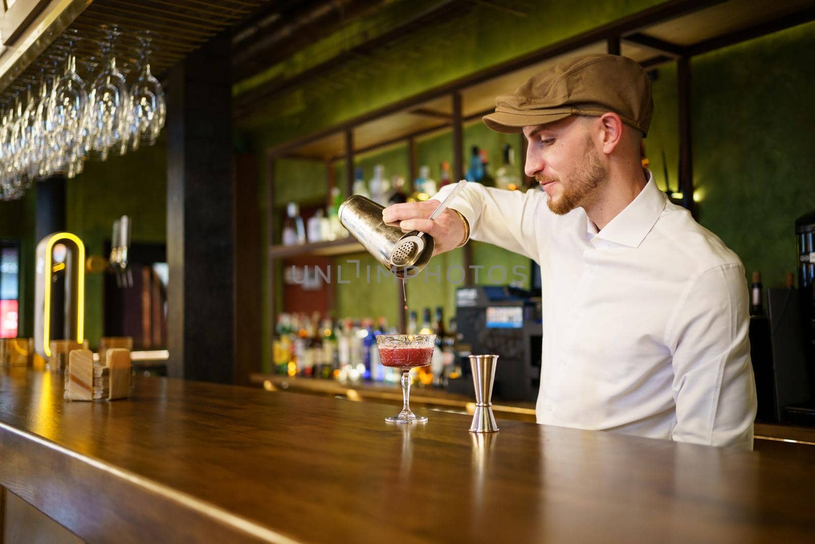 Bearded man in white shirt and cap filling glass with cocktail from shaker while standing behind counter and working in bar