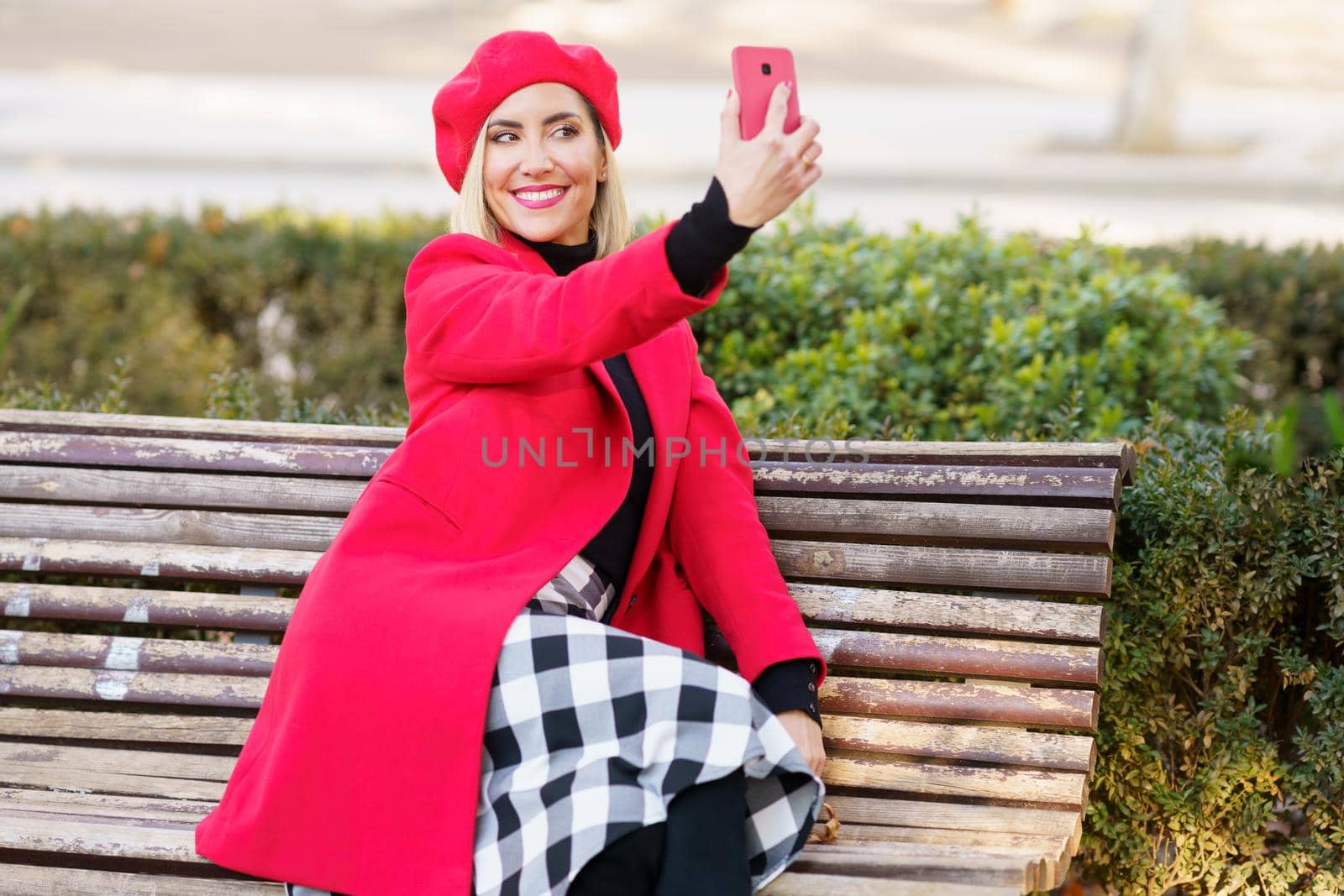 Delighted female in stylish red outfit taking self portrait on smartphone while sitting on wooden bench in park on autumn day
