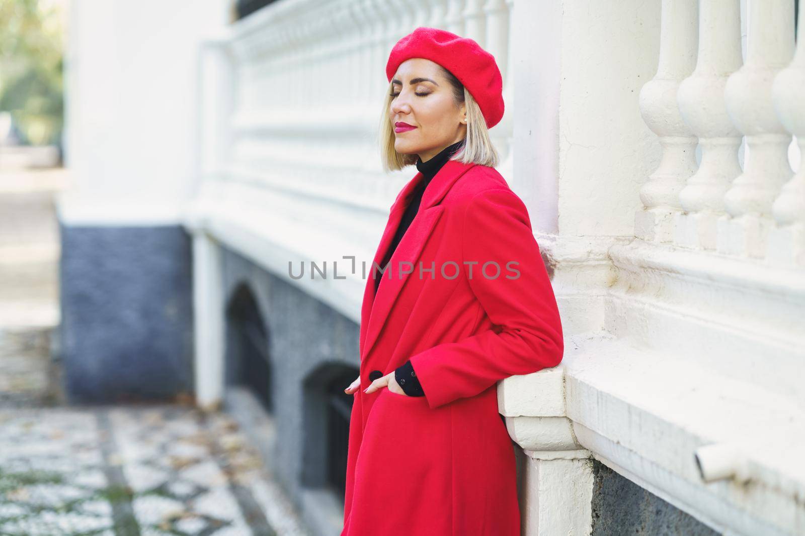 Side view of attractive female in red outfit with beret standing with hands in pockets near white fence on street