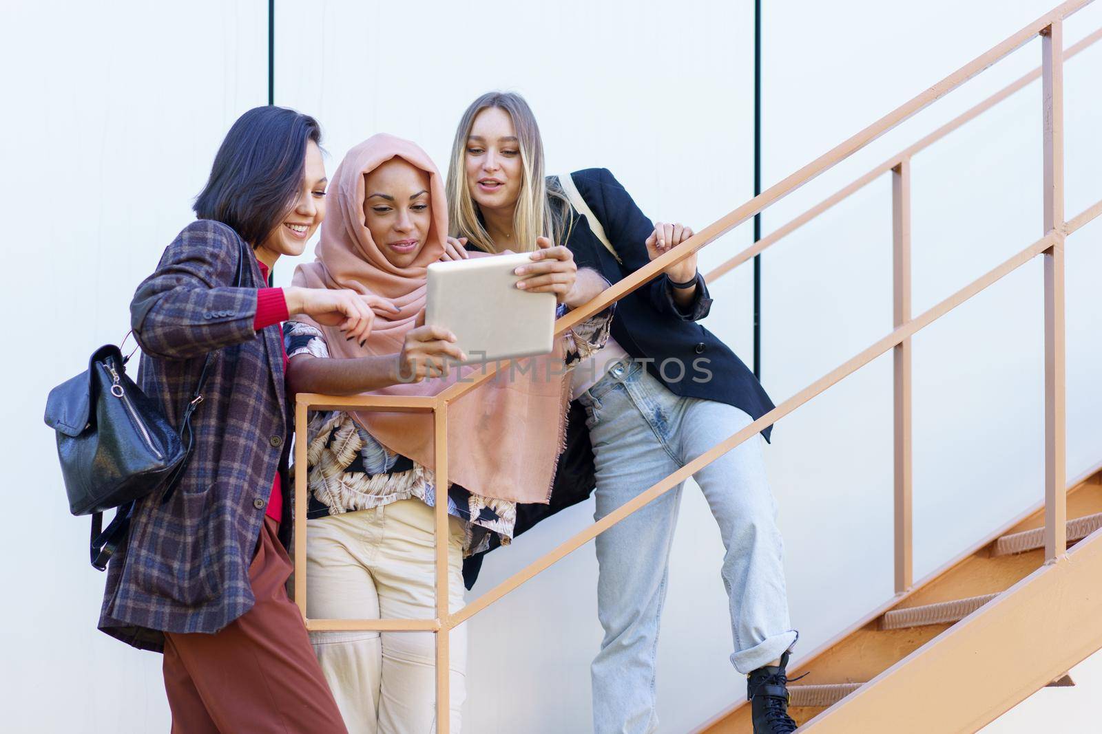 Confident young multiracial women smiling and sharing tablet standing on metal staircase on street