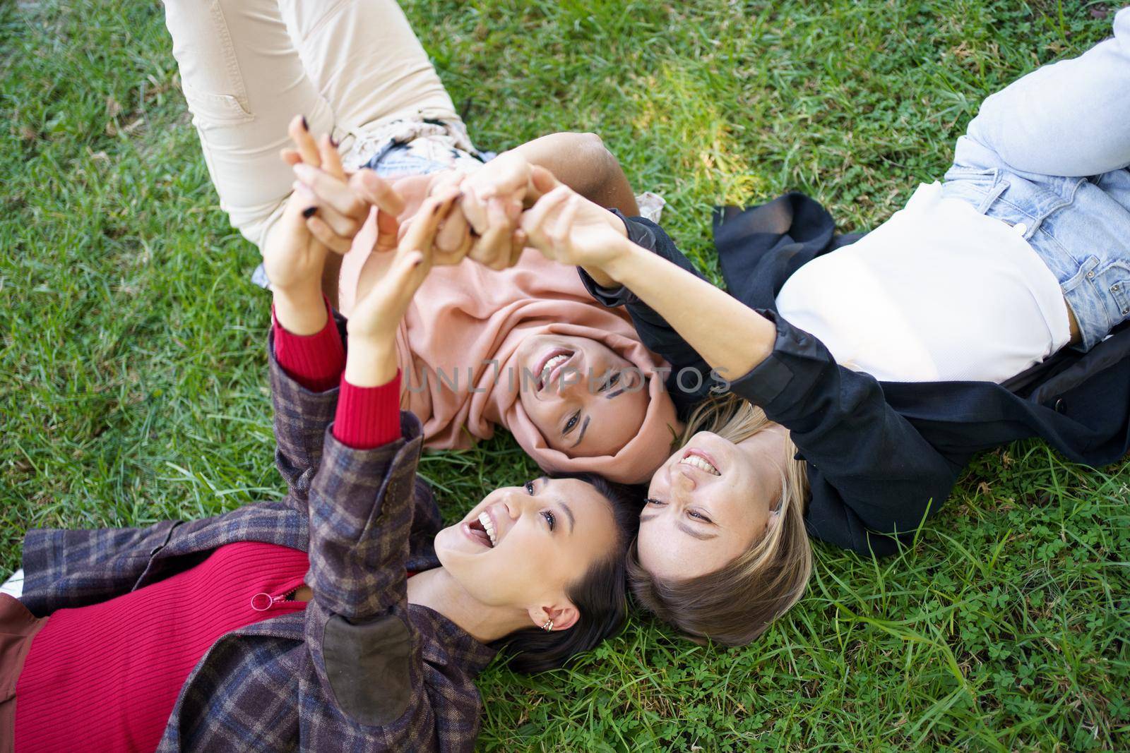 From above group of cheerful multiracial women holding hands together while lying on grassy lawn with raised arms in park