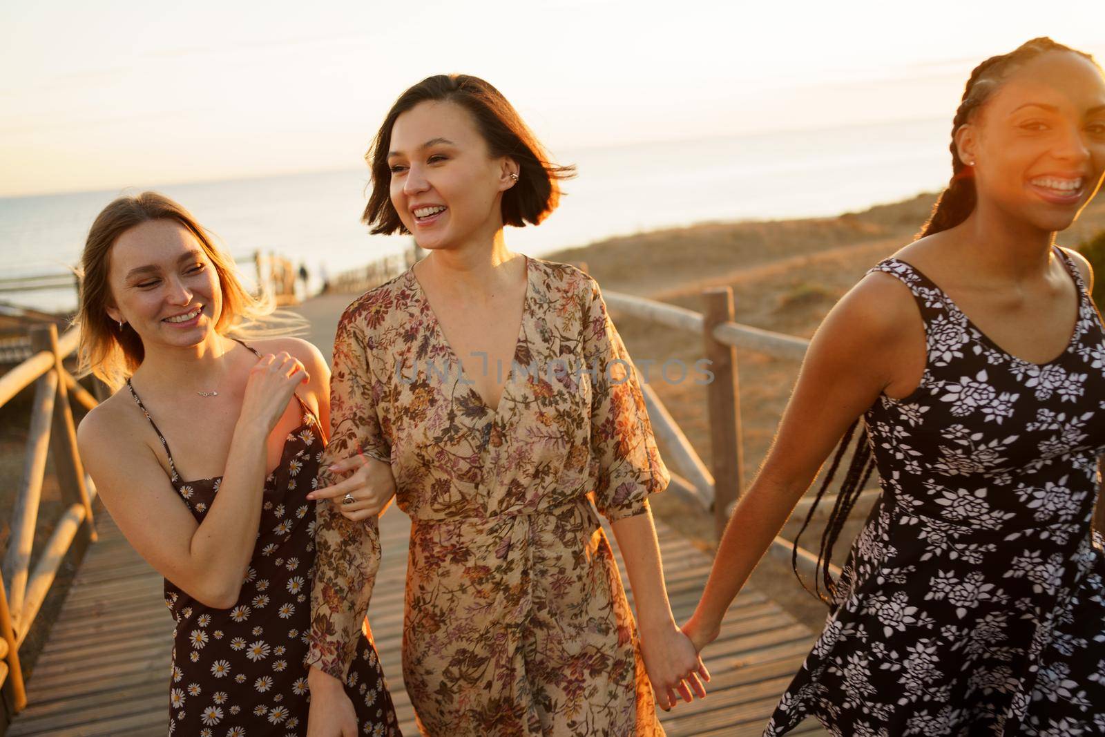 Optimistic multiracial female friends holding hands and walking together on wooden boardwalk while spending time together on beach against sea