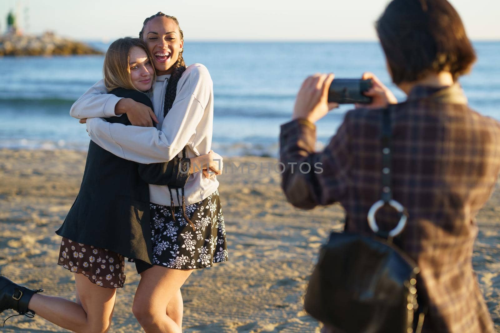 Faceless woman taking photo of hugging diverse girlfriends on seashore by javiindy