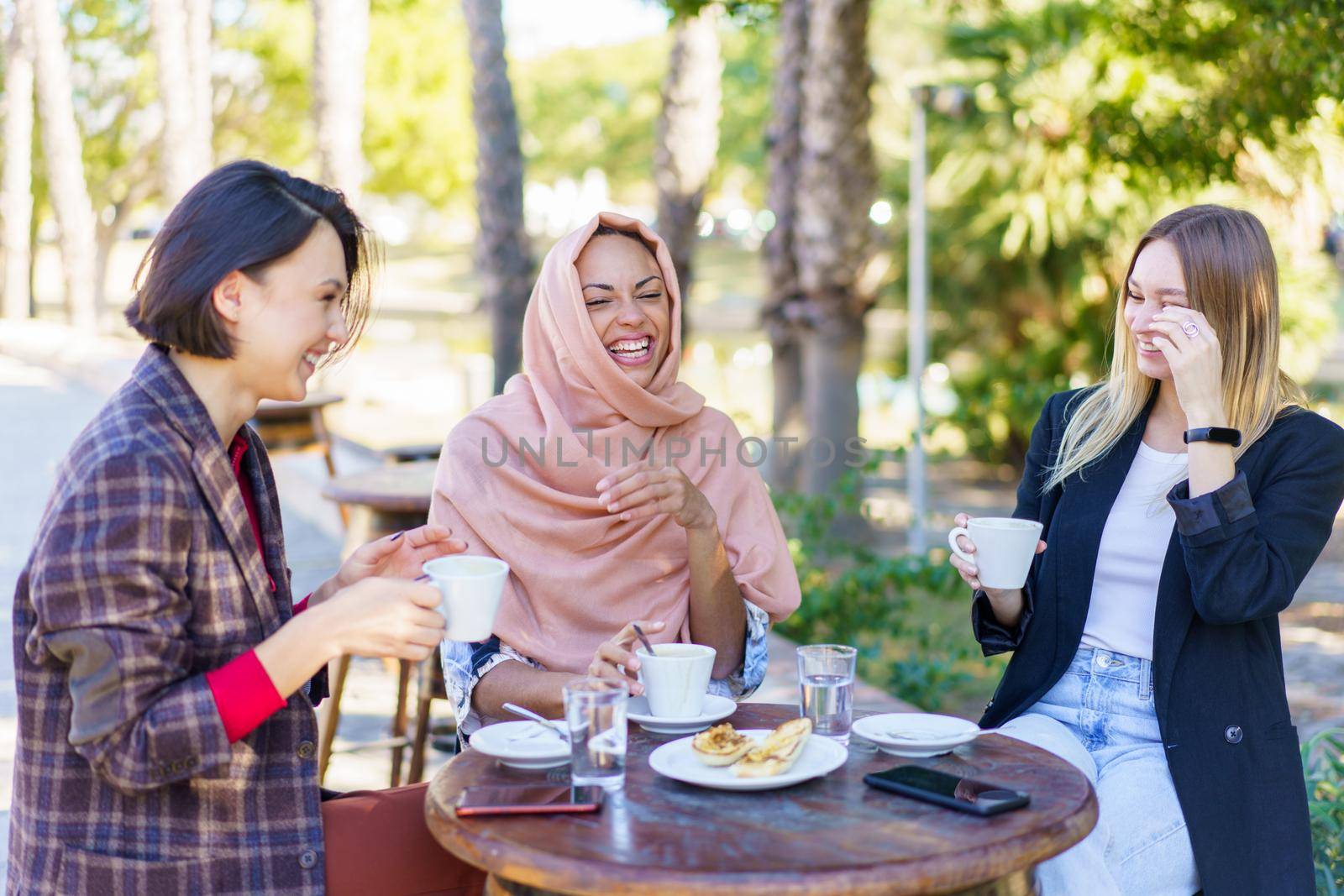Happy multiethnic female friends laughing while sitting at table with snack and coffee on terrace of cafeteria on summer day