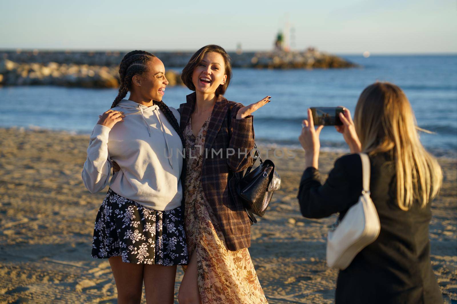 Blondie taking picture of excited multiracial girlfriends laughing joyfully and hugging each other on cellphone on sandy seashore