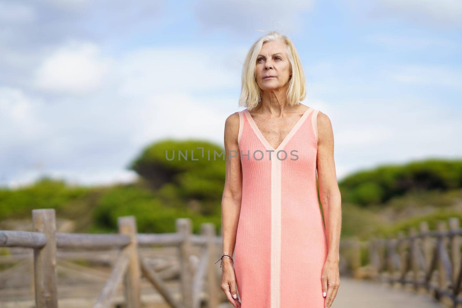 Beautiful mature woman walking along a wooden path near the beach., wearing a nice orange dress. Elderly female enjoying her retirement at a seaside retreat.