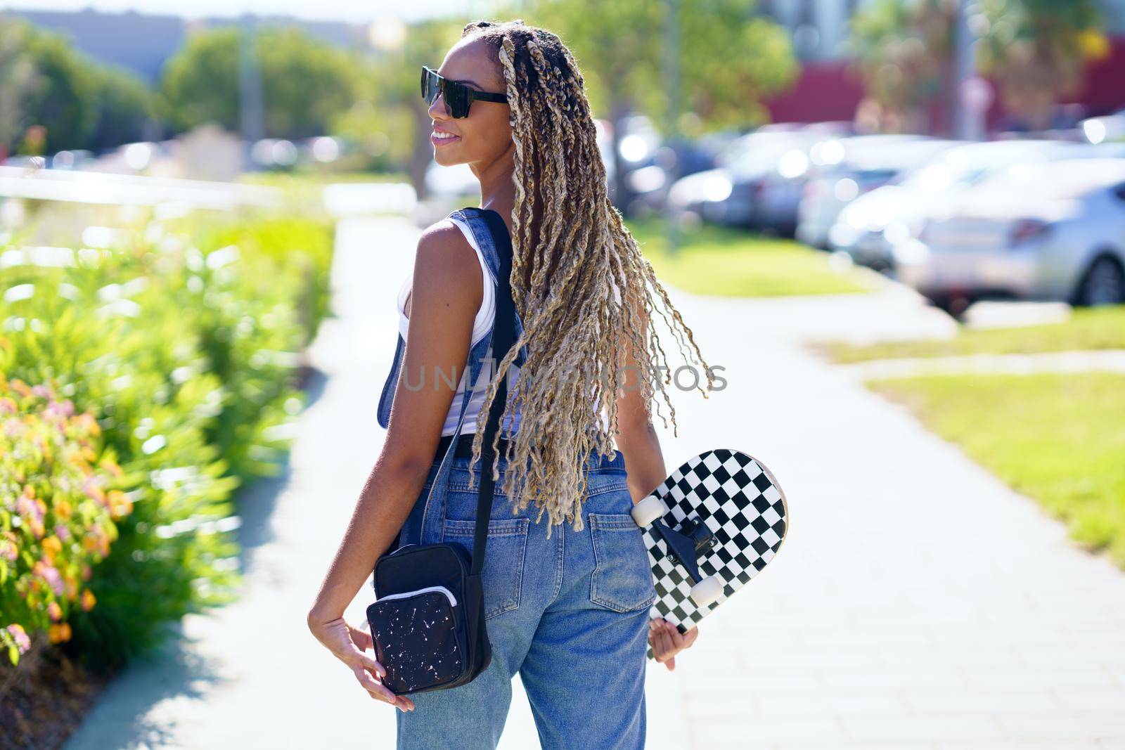 Back view of positive black female skater with braids and skateboard walking along street in summer and looking over shoulder