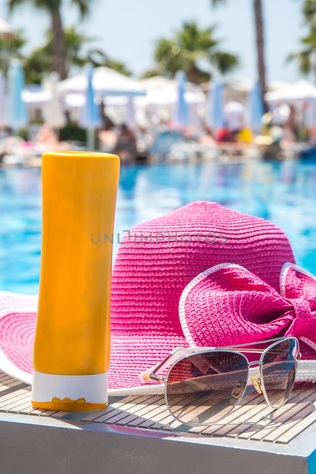 Bottle of sun cream, hat and sunglasses next to swimming pool in hotel by Mariakray