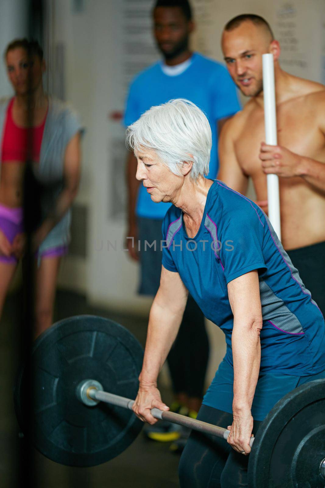 Shot of a senior woman lifting weights while a group of people in the background watch on.