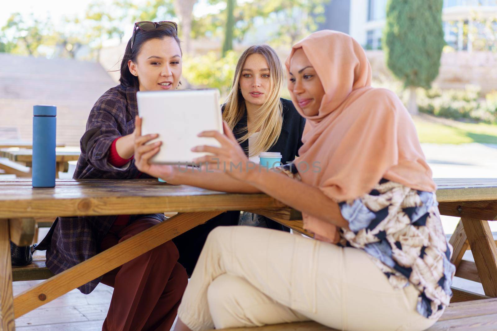 Group of positive young multiethnic women smiling and drinking coffee, to go while watching funny video on tablet, during break in outdoor cafe in park