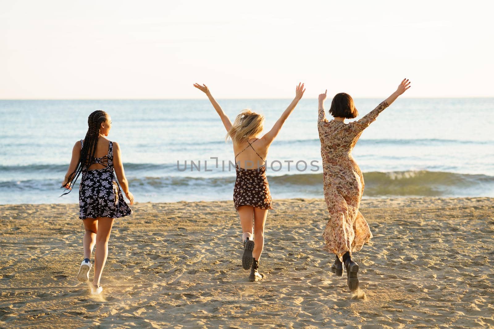 Full body back view of unrecognizable female friends in dresses running on sandy beach with raised arms near waving sea
