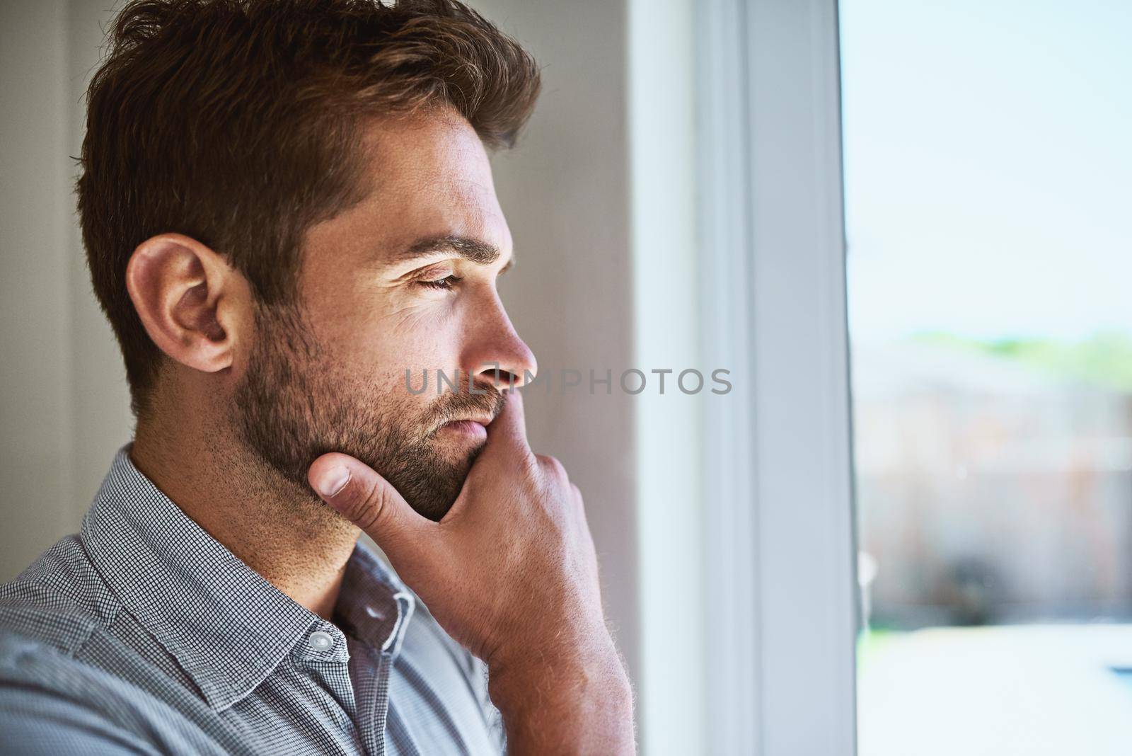 What can he be thinking about. Shot of a focused young man looking through a window while contemplating inside at home during the day. by YuriArcurs