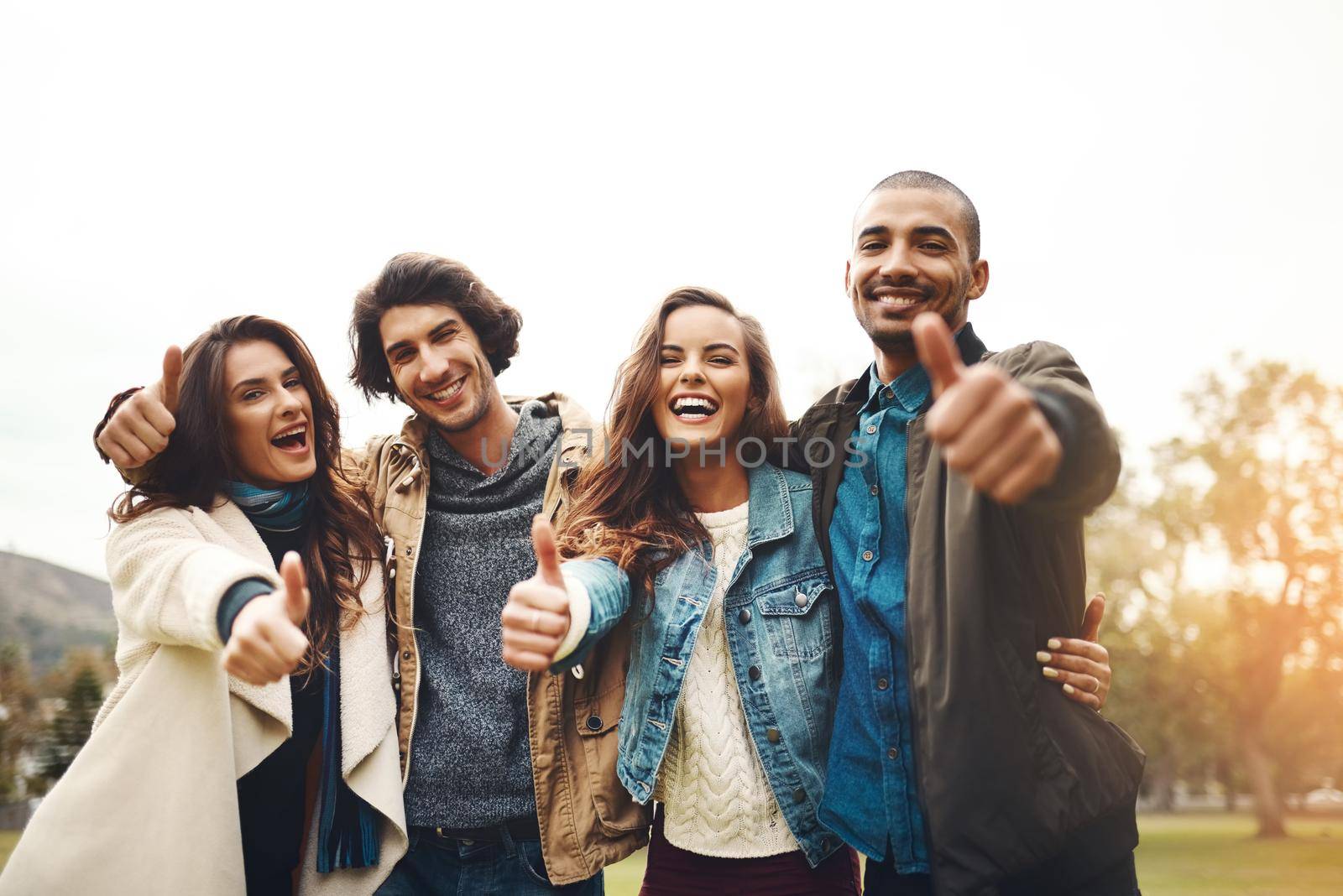 Portrait of a group of cheerful young friends huddled together while showing thumbs up outside during the day.