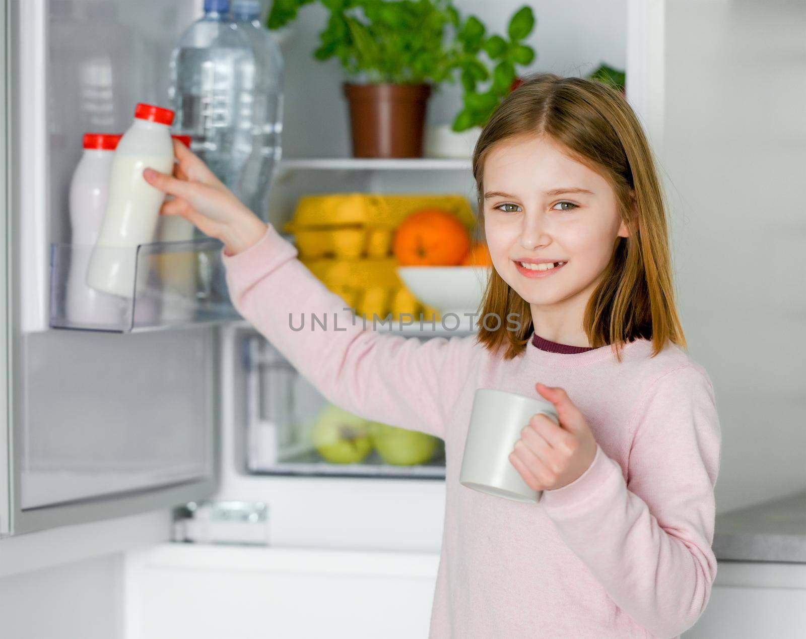 Pretty preteen girl taking yogurt from fridge and holding cup mug at kitchen at home. Beautiful female kid with milk products