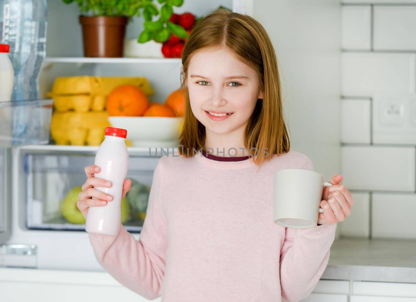 Pretty preteen girl holding yogurt and cup mug at kitchen at home and looking at camera. Beautiful female kid with milk products smiling