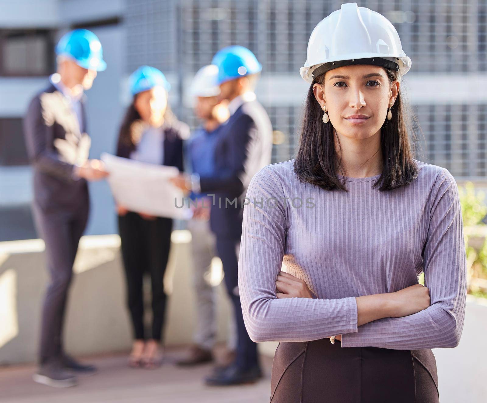 Shot of a young businesswoman standing with her arms folded and wearing a hardhat while her colleagues stand behind her.