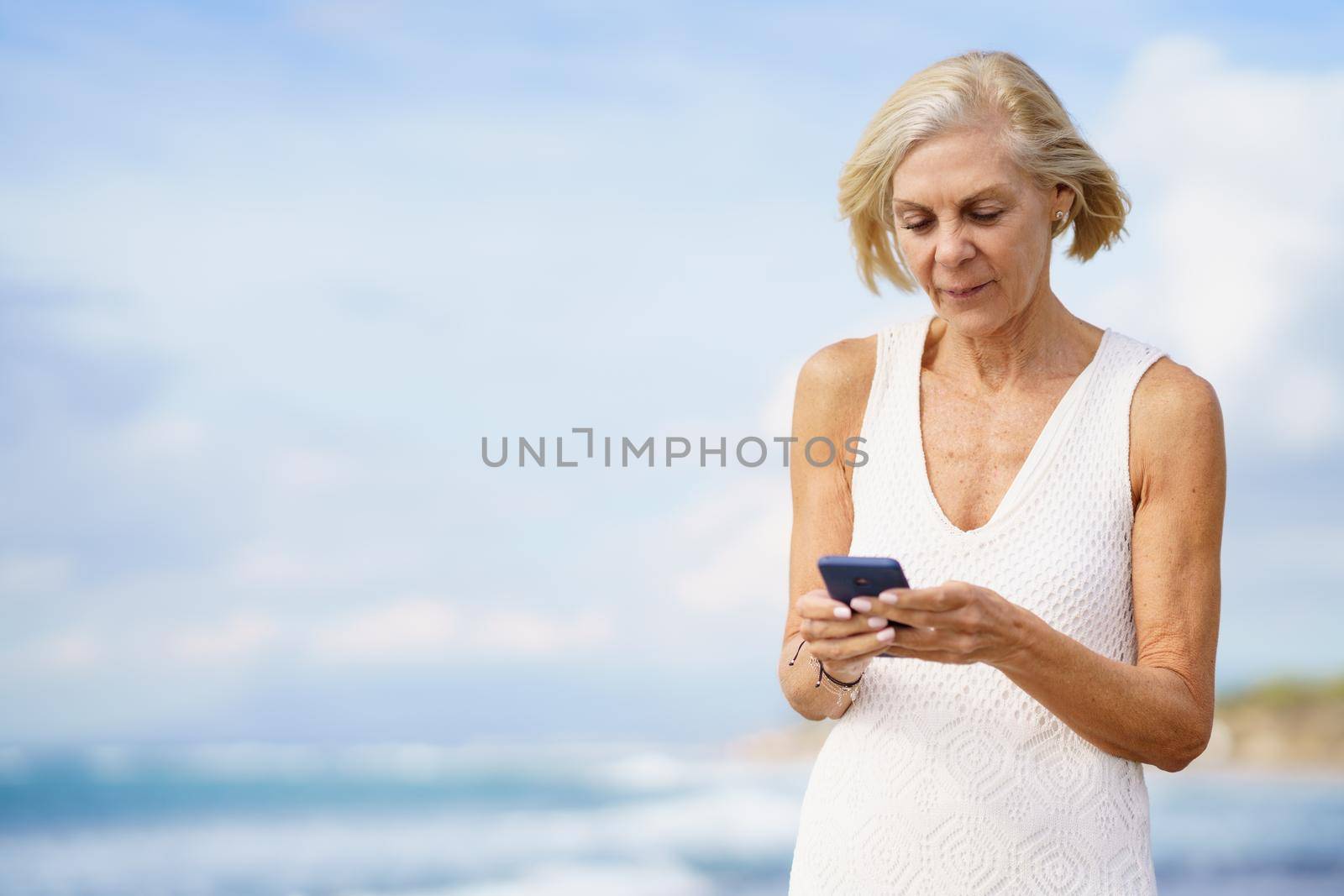 Mature woman walking on the beach using a smartphone. Elderly female enjoying her retirement at a seaside location.
