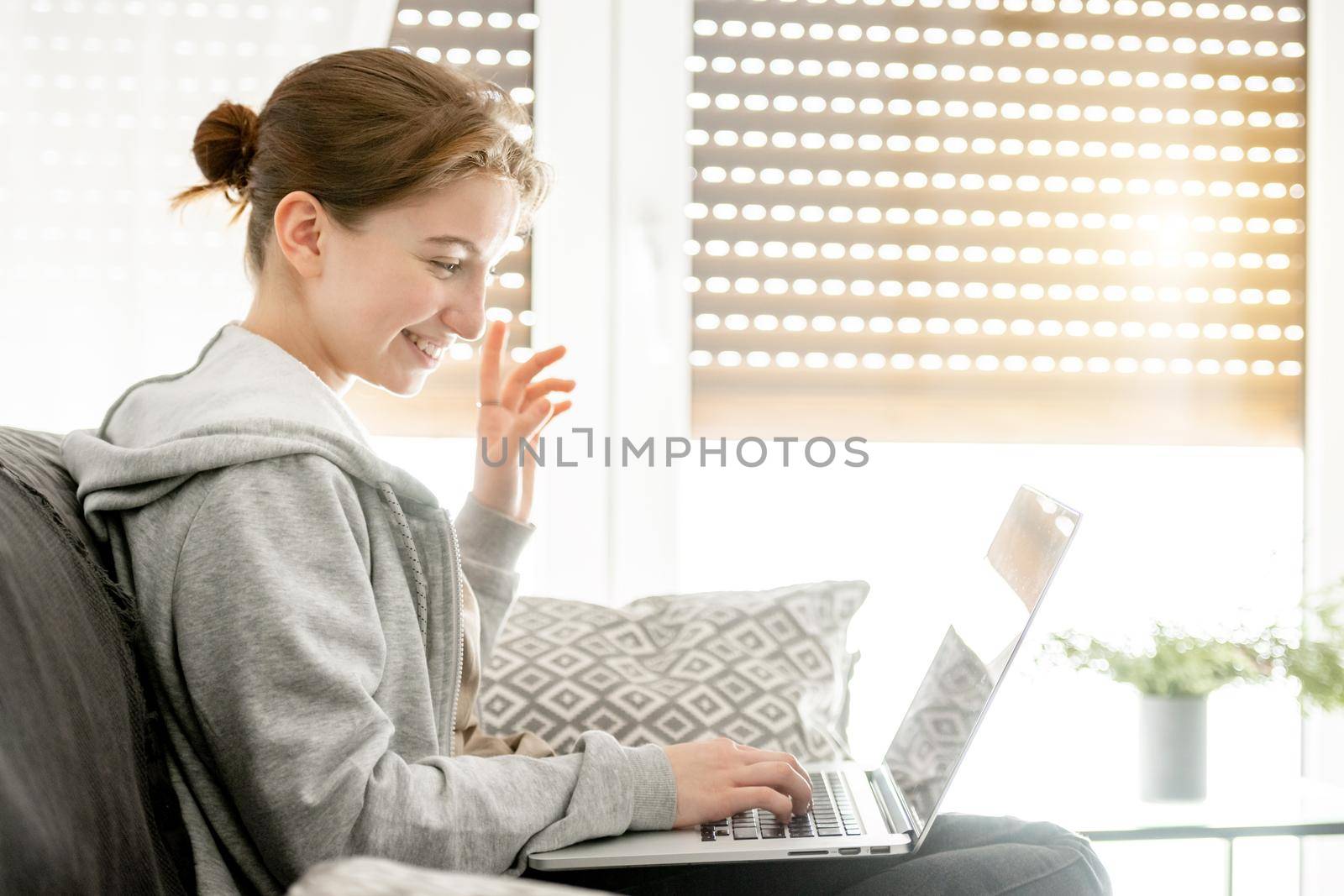 Smiling girl sitting on sofa with laptop and studying online with remote education programm in covid pandemic time. Happy teenager spends time at computer with classmates friends