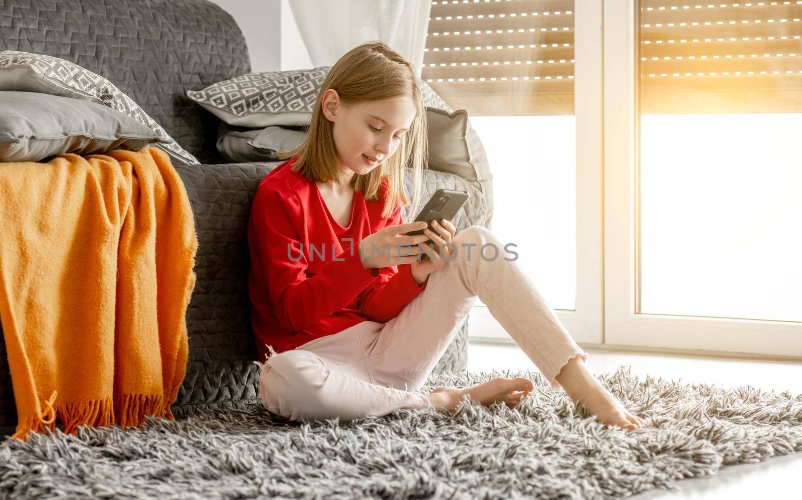 Child girl holds smartphone in her hand and smiles chatting with friends. Cheerful kid spends time with cellphone sitting on floor near sofa by window.