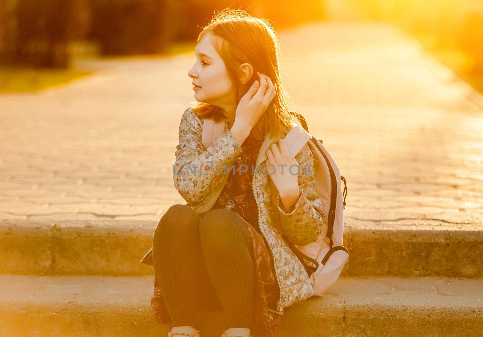 Preteen girl child with backpack sitting at street and looking back in amazing sunset light. Pretty schoolgirl going home after class