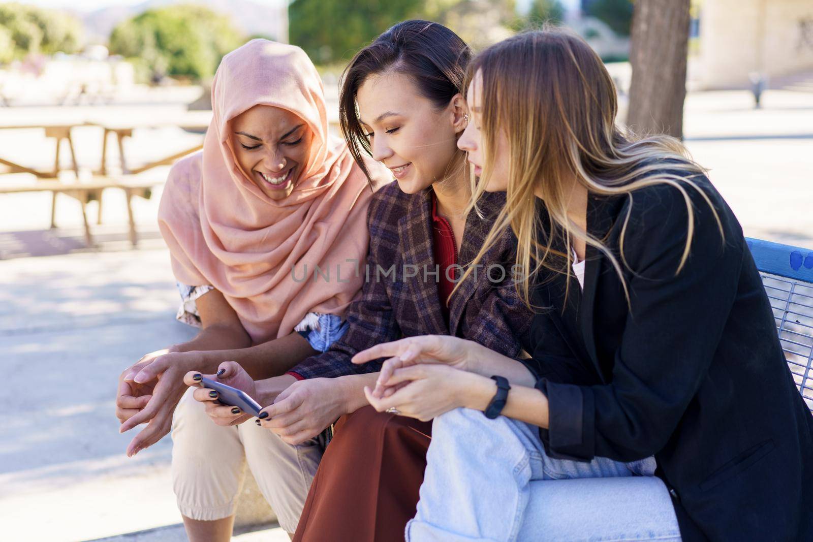 Positive multiethnic girlfriends sharing smartphone while resting on bench in park by javiindy