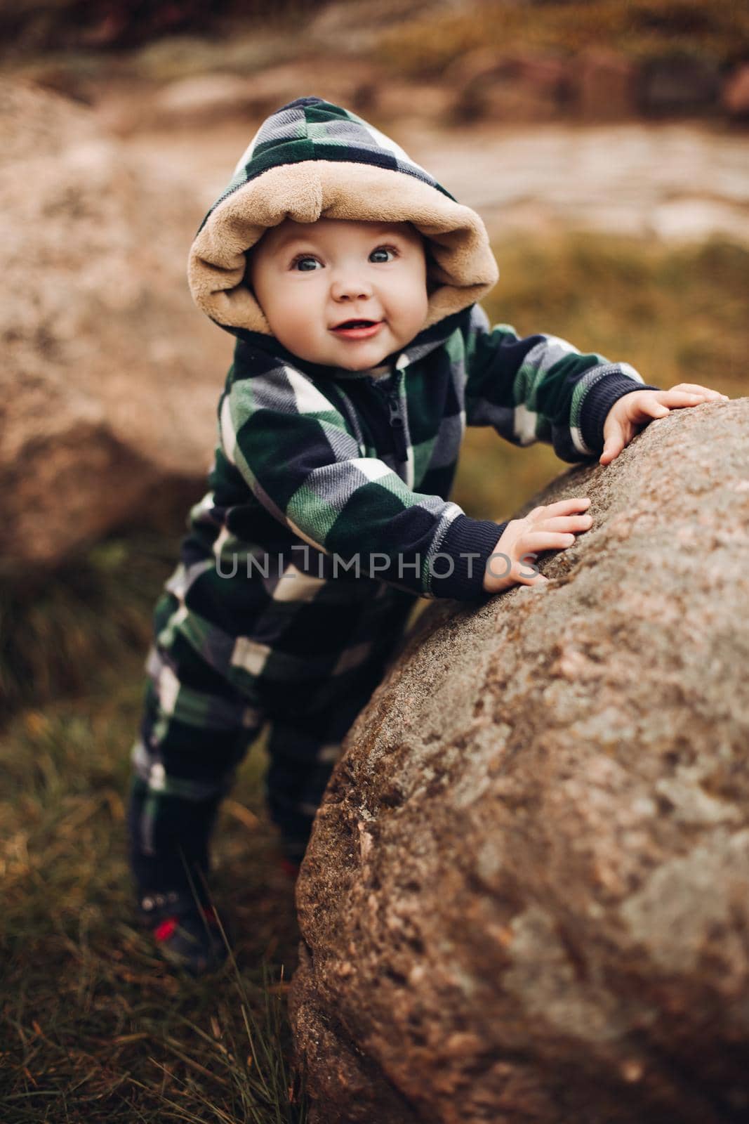 Portrait of cute baby in checked green, blue and black overall with hood sitting on the ground covered with bright dry foliage on autumn day. Fair haired baby in overall on vivid colorful leaves in the autumnal park or forest.