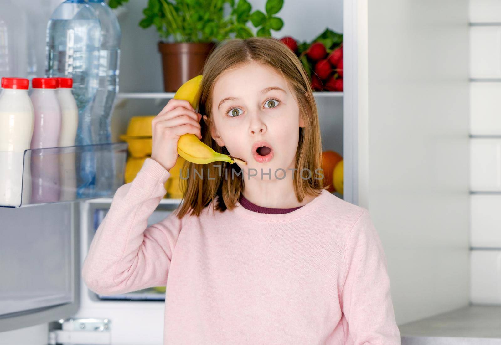Pretty preteen girl holding banana as smartphone at kitchen and playing with fruit. Beautiful female kid happy at home