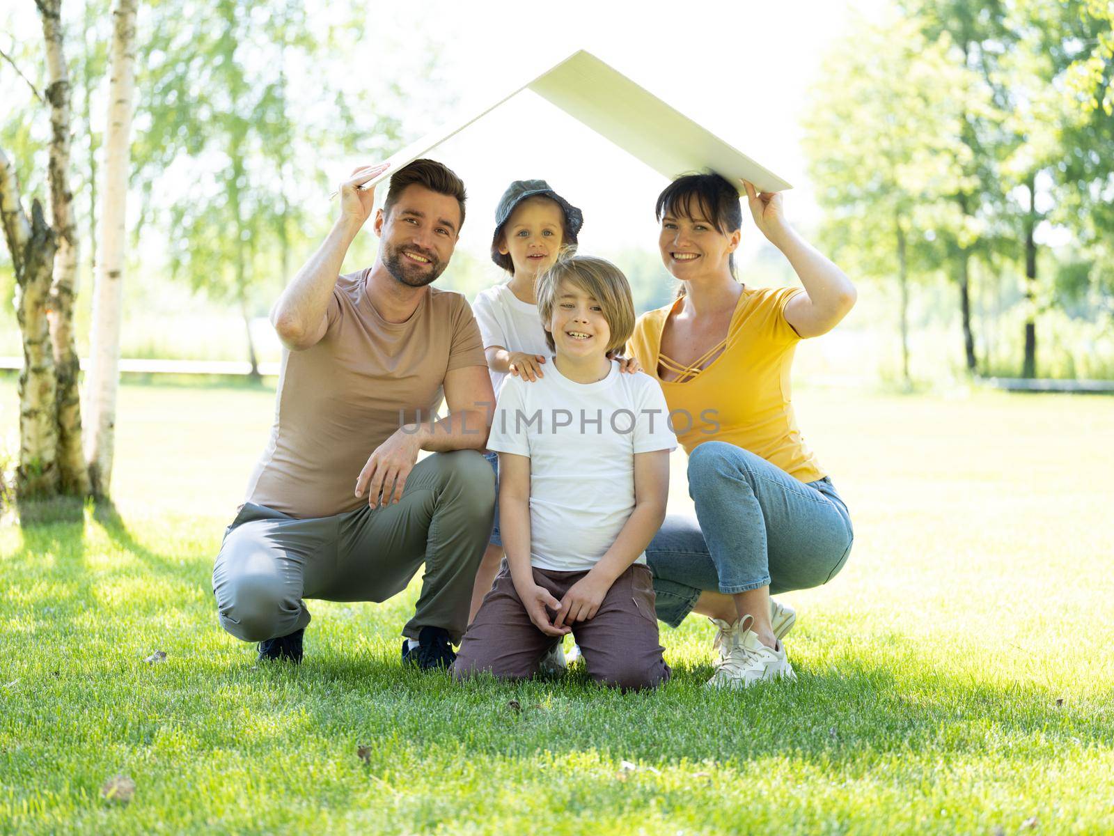 Concept of mortgage and housing for young families. Mother father and two children sitting on grass and holding roof symbol