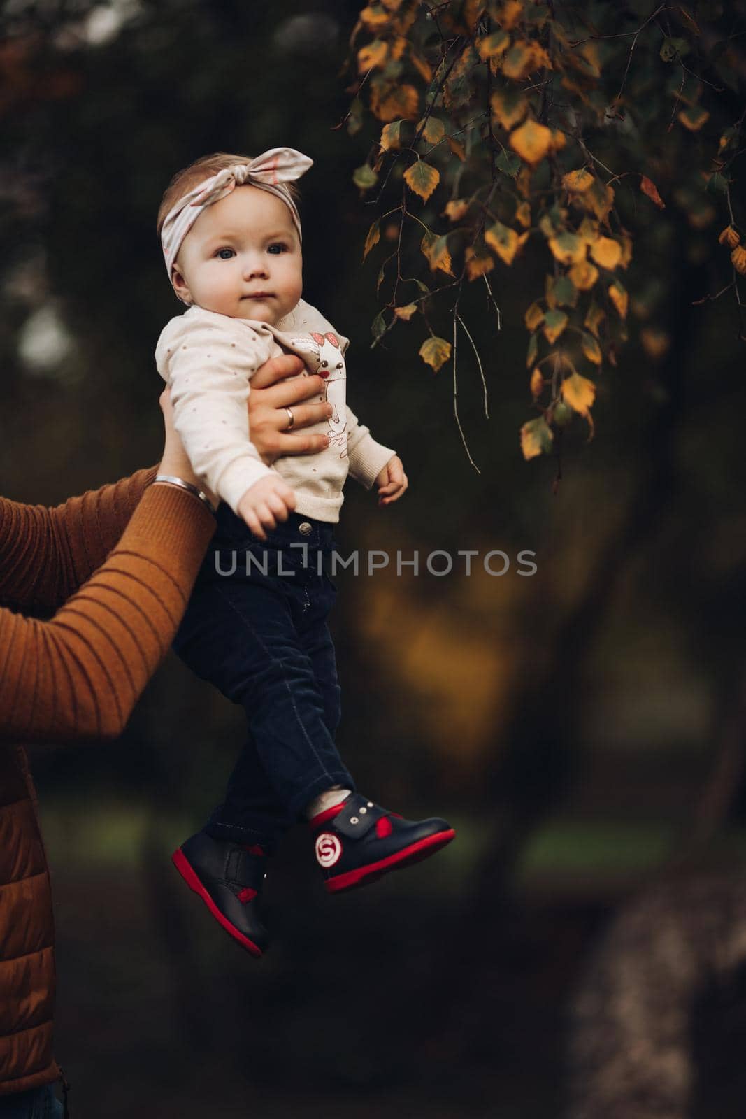 Portrait of lovely little baby girl wearing cute jersey with bunny on it and beige patterned bow headband. She is smiling at camera while parents holding her arms. Weekend in the park in autumn. Baby doing her first steps.