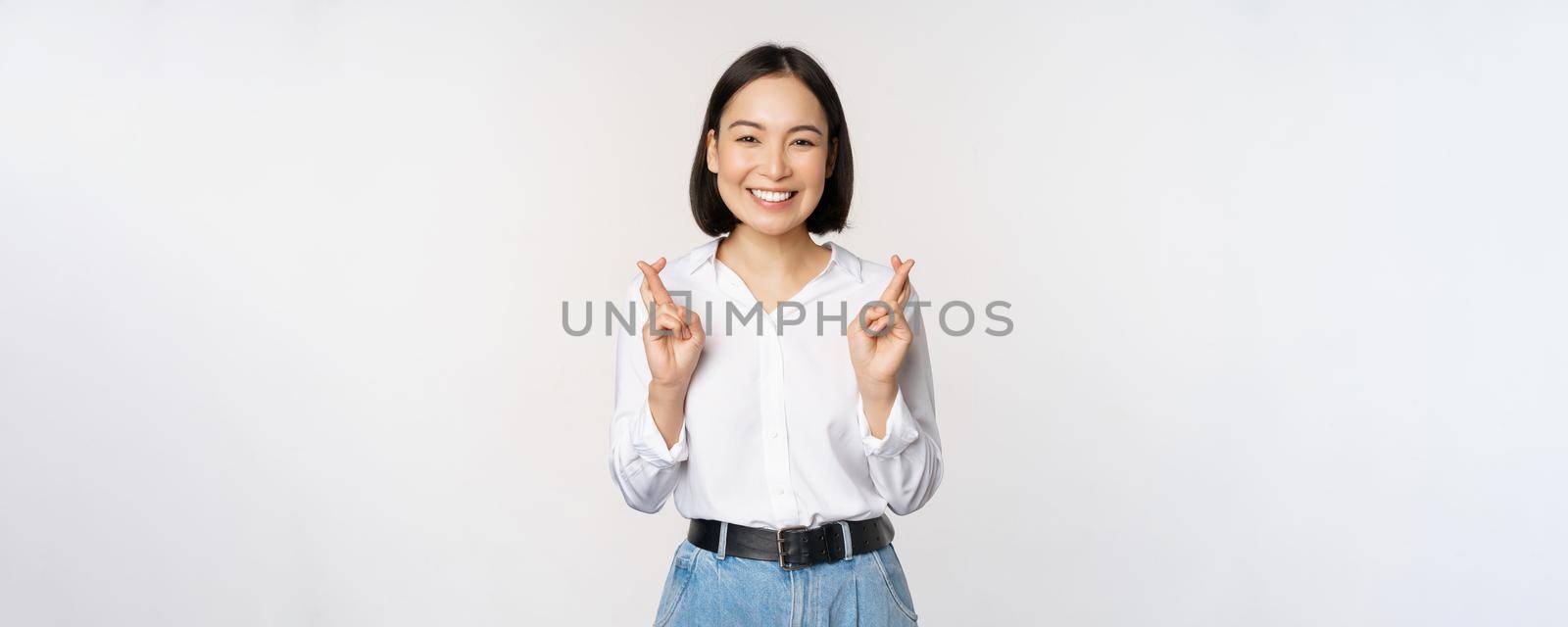 Portrait of young korean woman, asian girl cross fingers and praying, making wish, anticipating, waiting for results, standing over white background.