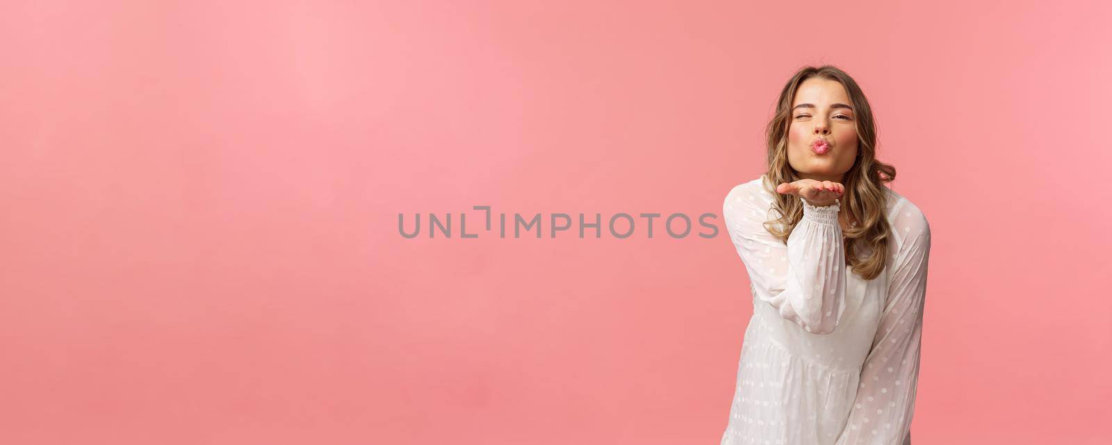 Beauty, fashion and women concept. Portrait of romantic tender, young blond woman in white trendy dress, sending air kiss at camera with sassy flirty smile, hold hand near lips, pink background by Benzoix