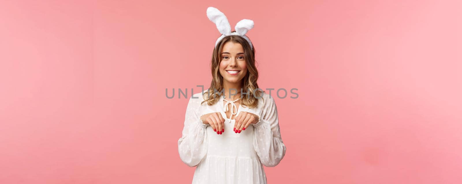 Holidays, spring and party concept. Portrait of cute and tender, lovely blond girl imitating bunny, holding hands like paws and wearing rabbit ears, smiling camera, pink background.