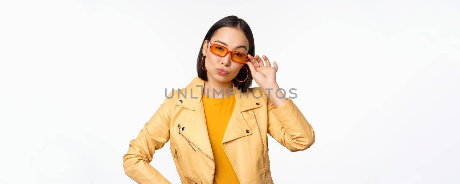 Stylish asian female model, wears sunglasses, looks confident and cool, stands over white background.